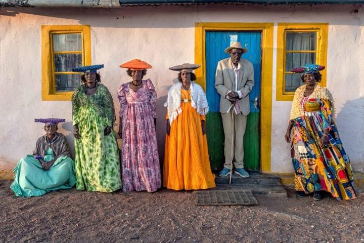 Farbfoto von heute: Herero-Männer und -Frauen in traditioneller Tracht vor einem Haus
