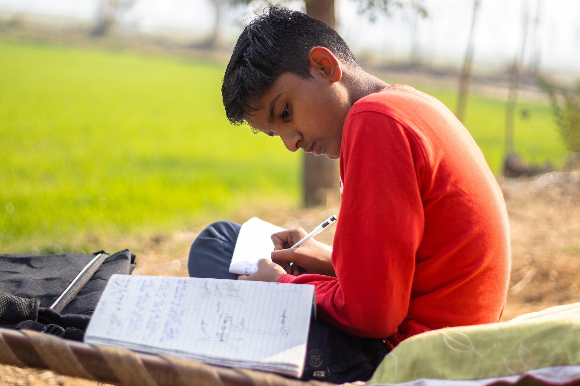 A boy sitting outdoors on a cot, writing in a notebook, with fields in the background